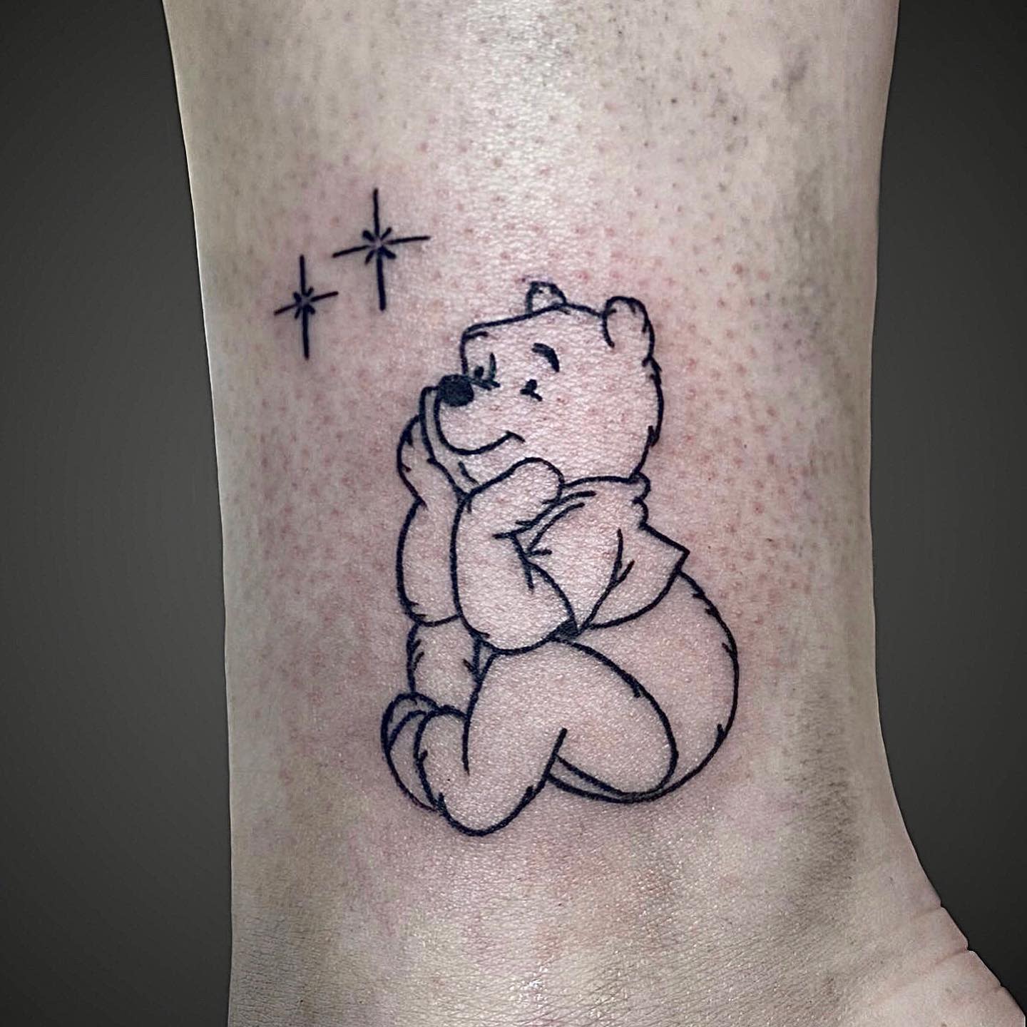 Outline tattoo of Winnie the Pooh sitting with a content expression and two star symbols above him on a person's leg